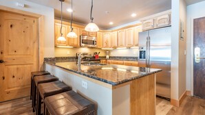 A kitchen with wooden cabinets, a stainless steel refrigerator, a granite countertop, and pendant lighting.