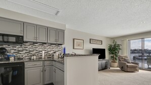 Una cucina moderna con mobili grigi, un forno a microonde nero e uno schienale rivestito di piastrelle. C'è un'area pranzo con una TV a schermo piatto e una poltrona comoda. Un'ampia finestra offre vista su un corpo d'acqua.