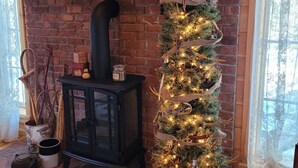 Een knus vertrek met een metselwerk openhaard, een houten plankje met diverse decoraties, een zwarte kachel en een kerstboom.