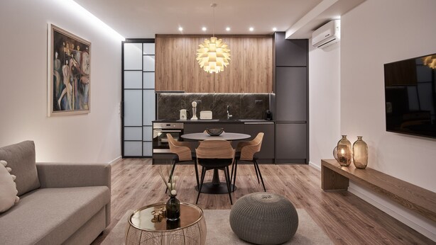 Uma sala de estar moderna com sofá, mesa de centro e um banquinho, uma área de jantar com mesa e cadeiras, e uma cozinha com ilha central, luminária pendente e eletrodomésticos embutidos.