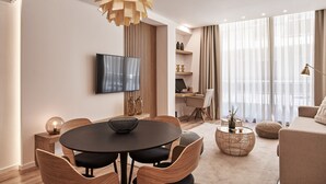 Uma sala de estar moderna com uma mesa de jantar redonda, cadeiras de madeira, TV na parede, escrivaninha com computador e uma grande janela com cortinas leves.