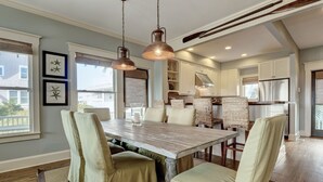 A dining area with a large wooden table, cushioned chairs, pendant lighting, and a view of the kitchen in the background. - Executive seaside cottage with detached Crofter Cottage. (Bald Head Island)