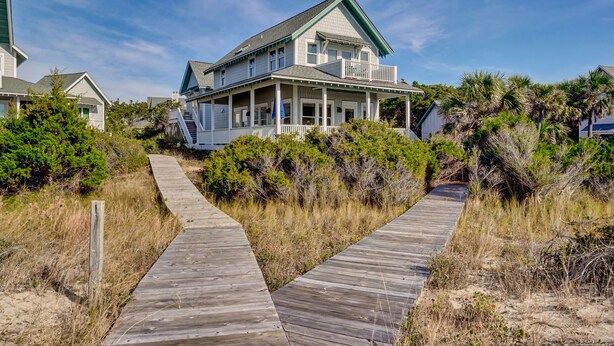 A coastal house with a wooden deck leading to the entrance, surrounded by dense vegetation and other houses. - Executive seaside cottage with detached Crofter Cottage. (Bald Head Island)