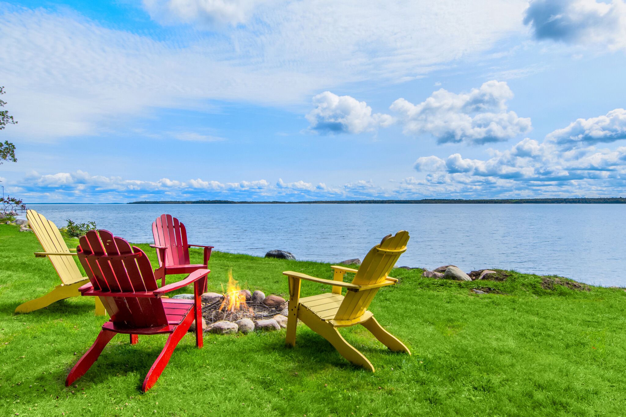 A lakeside view with two Adirondack chairs and a campfire.