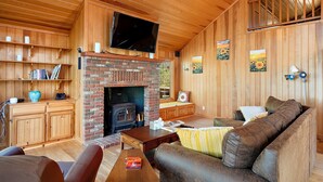 A cozy living room with a brick fireplace, wooden walls, a flat-screen TV, and comfortable seating.
