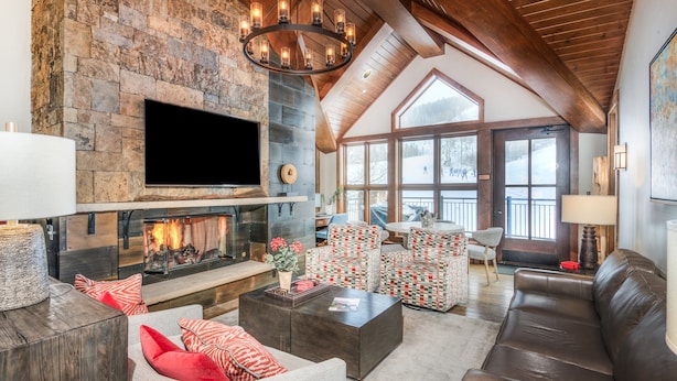 A spacious living room with a stone fireplace, a large TV, comfortable seating, and a high ceiling with wooden beams.
