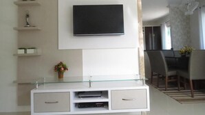 Una sala de estar moderna con un televisor de pantalla plana montado, un mueble de entretenimiento con cubierta de vidrio y un comedor con candelabro.