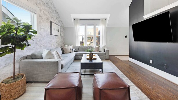 A modern living room with a grey sectional sofa, a coffee table, and leather ottomans. A large flat-screen TV is mounted on the wall, and a potted plant is visible.