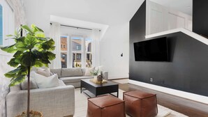 A modern living room with a grey sofa, a black coffee table, and two brown ottomans. A large TV is mounted on the wall, and a potted plant is in the corner.