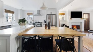 A modern kitchen with white cabinetry, a large wooden island, black chairs, and a fireplace.