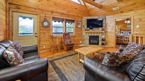A cozy log cabin interior with a leather sofa, a wooden coffee table, a stone fireplace, and a television mounted on the wall. - Epic Views, Perfect Location, Fast WiFi (Gatlinburg)