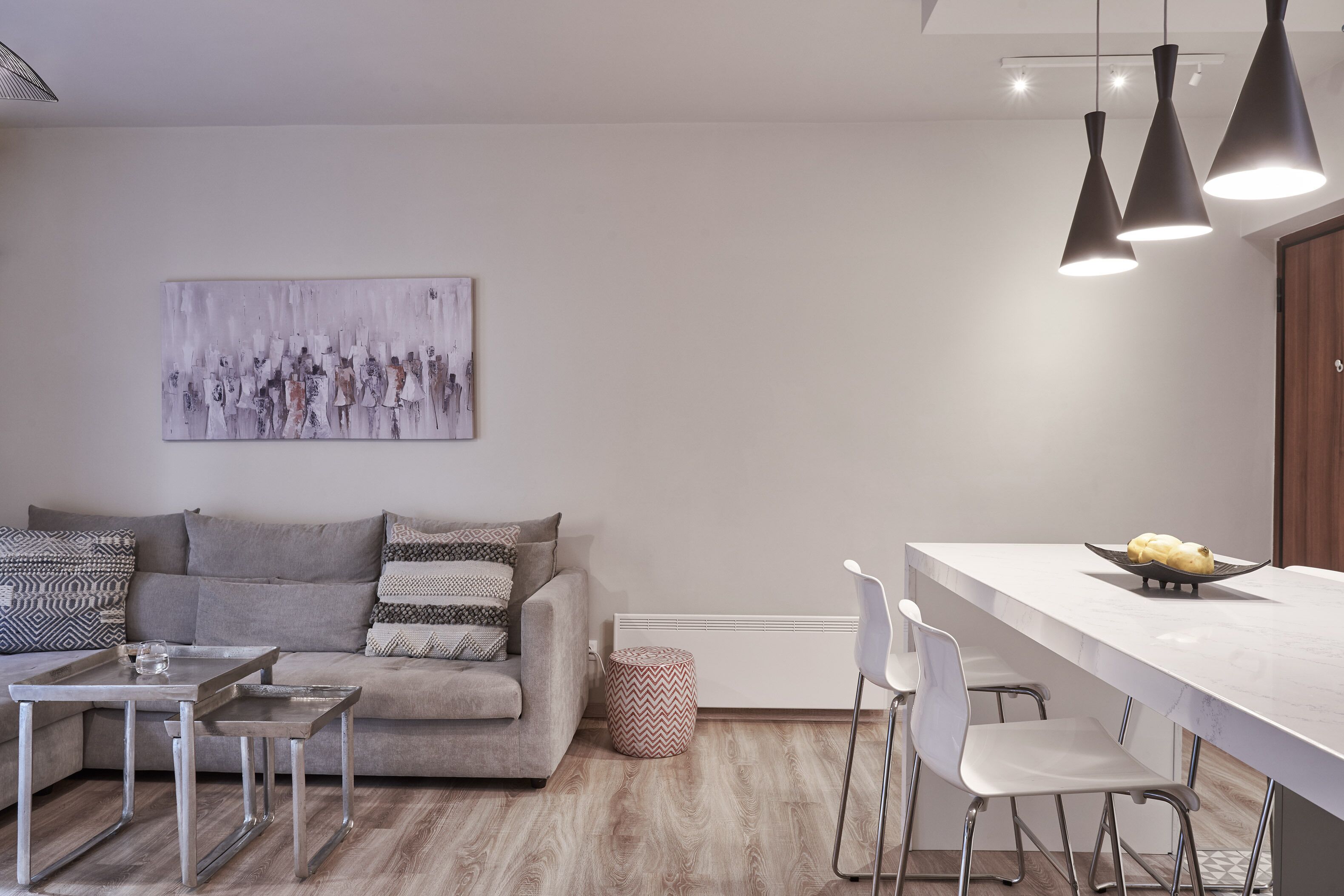 A modern living room with a grey sofa, a small dining table, and a kitchen area with a white countertop and bar stools.