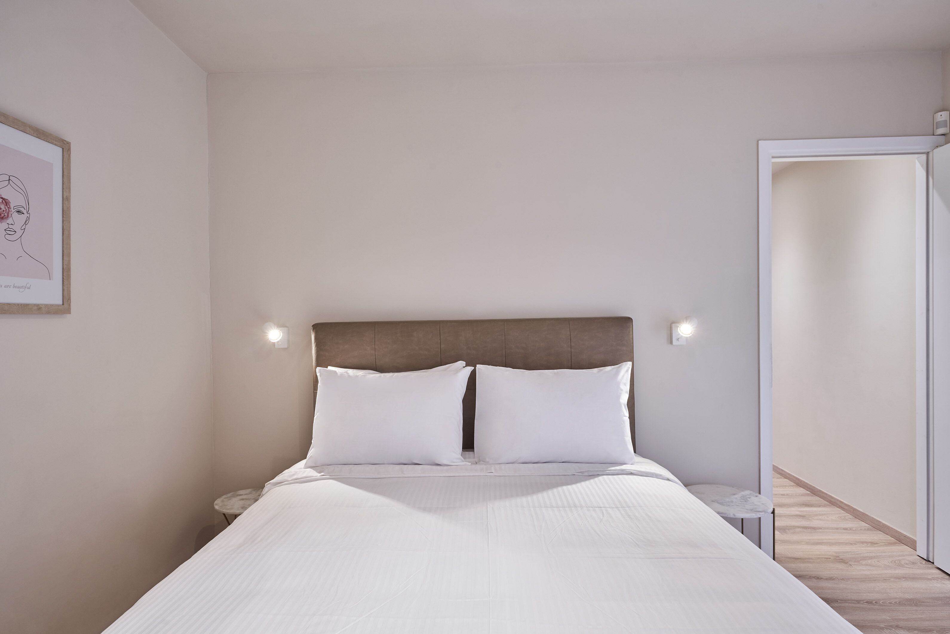 Una cama bien hecha con ropa blanca y un cabecero de madera, dos mesitas de noche con lámparas y un cuadro en la pared.