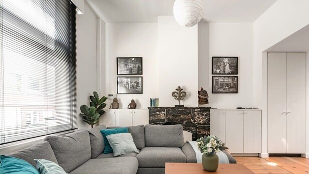 A modern living room with a grey sectional sofa, a wooden coffee table, and a large window with blinds. There are two framed pictures on the wall and a white cabinet in the background. - Urban Comfort: Central 2-BR Townhouse for 6 (Gent)