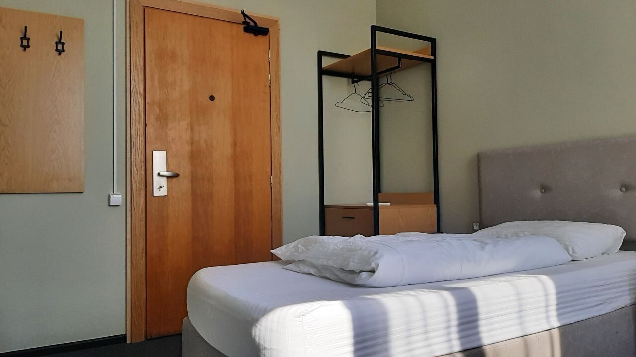 A bedroom with a wooden door, a bed with white linens, a hanging shoe rack, and a mirror.