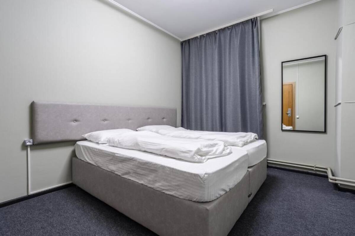 A hotel room with a single bed, a grey headboard, a grey curtain, a mirror, and a grey carpet.