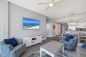 Un salon moderne avec un grand téléviseur, des canapés à motifs, une table basse et une vue sur la plage par la fenêtre.