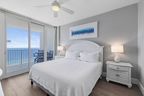 Een slaapkamer met een groot bed, een plafondventilator, een nachtkastje met lamp, een schilderij aan de muur en een balkon met uitzicht op de oceaan.
