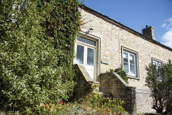 A stone building with two windows and a door, surrounded by greenery.