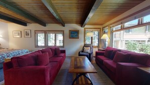 Ein Zimmer mit zwei roten Sofas, einem Holztisch und einem Bett mit gemusterten Betttextilien. Es gibt Fenster mit Vorhängen, eine Lampe und gerahmte Bilder an den Wänden.