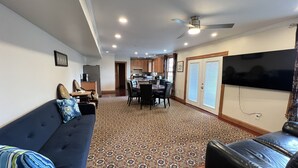 A spacious living room with a dark blue sofa, a patterned rug, and a leather chair. A dining area with a table and chairs is visible in the background. - 3BR/3BA + 3 Parking — Harvard/MIT, Red & Green Lines (Cambridge)