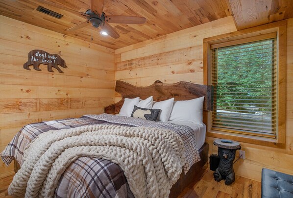 Une chambre confortable, aménagée dans un chalet en bois, avec un lit, un ventilateur de plafond, une fenêtre avec des stores et une décoration représentant un ours sur le mur.