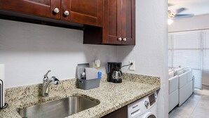 Cocina con encimeras de granito, pileta de acero inoxidable, muebles de madera, cafetera y lavadora.