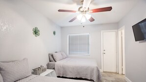 Un dormitorio con dos camas, ventilador de techo, ventana con persianas, televisor montado en la pared y una puerta.