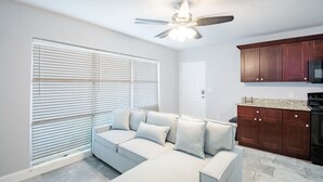 Una sala de estar moderna con un sofá seccional, un ventilador de techo y una cocina con armarios de madera y un microondas.