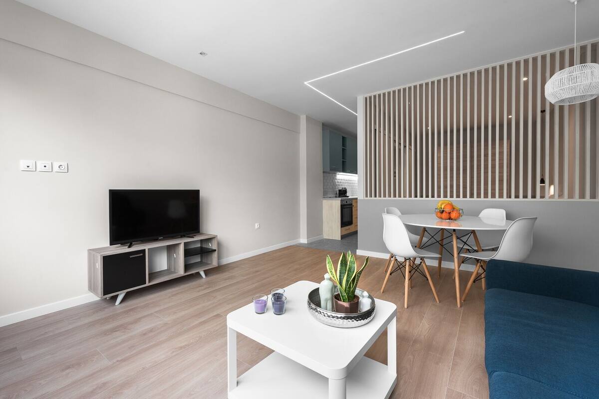 A modern living room with a flat-screen TV, a white coffee table with decorative items, a blue sofa, and a dining area with a white table and chairs.