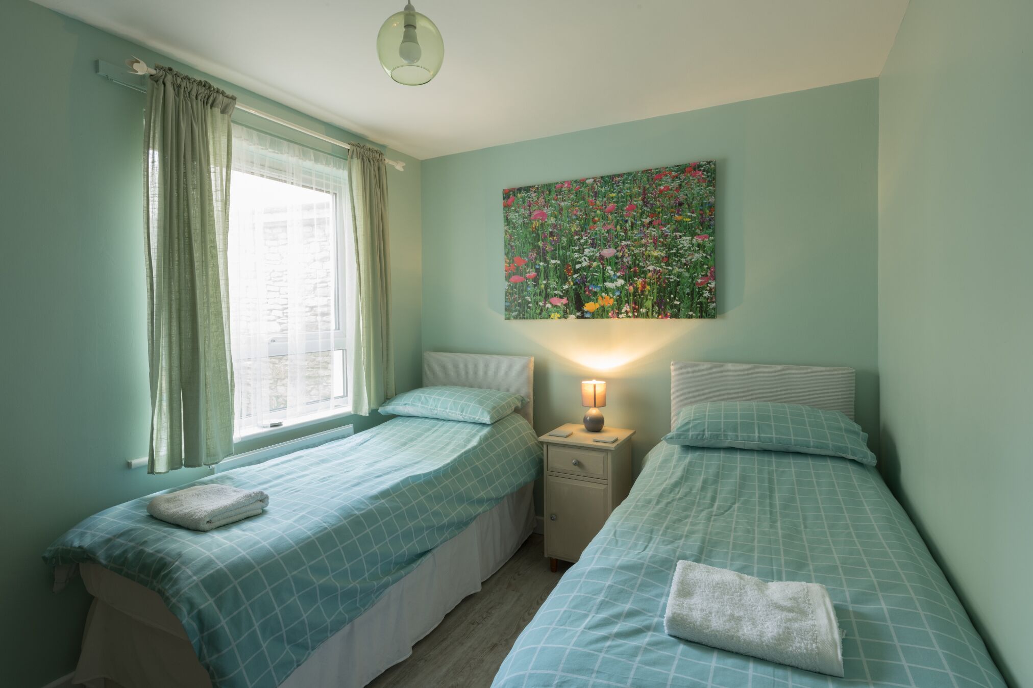 A bedroom with two beds, a nightstand, a lamp, a window with curtains, and a floral painting on the wall.