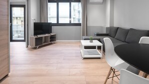 A modern living room with a flat-screen TV, grey sofa, white chairs, and a small table with a plant. - New Yorker Nilie Hospitality MGMT (Thessaloniki)