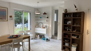 Ein Essbereich mit Holztisch und weißen Stühlen, eine Küche mit offenen Regalen und Blick ins Freie.
