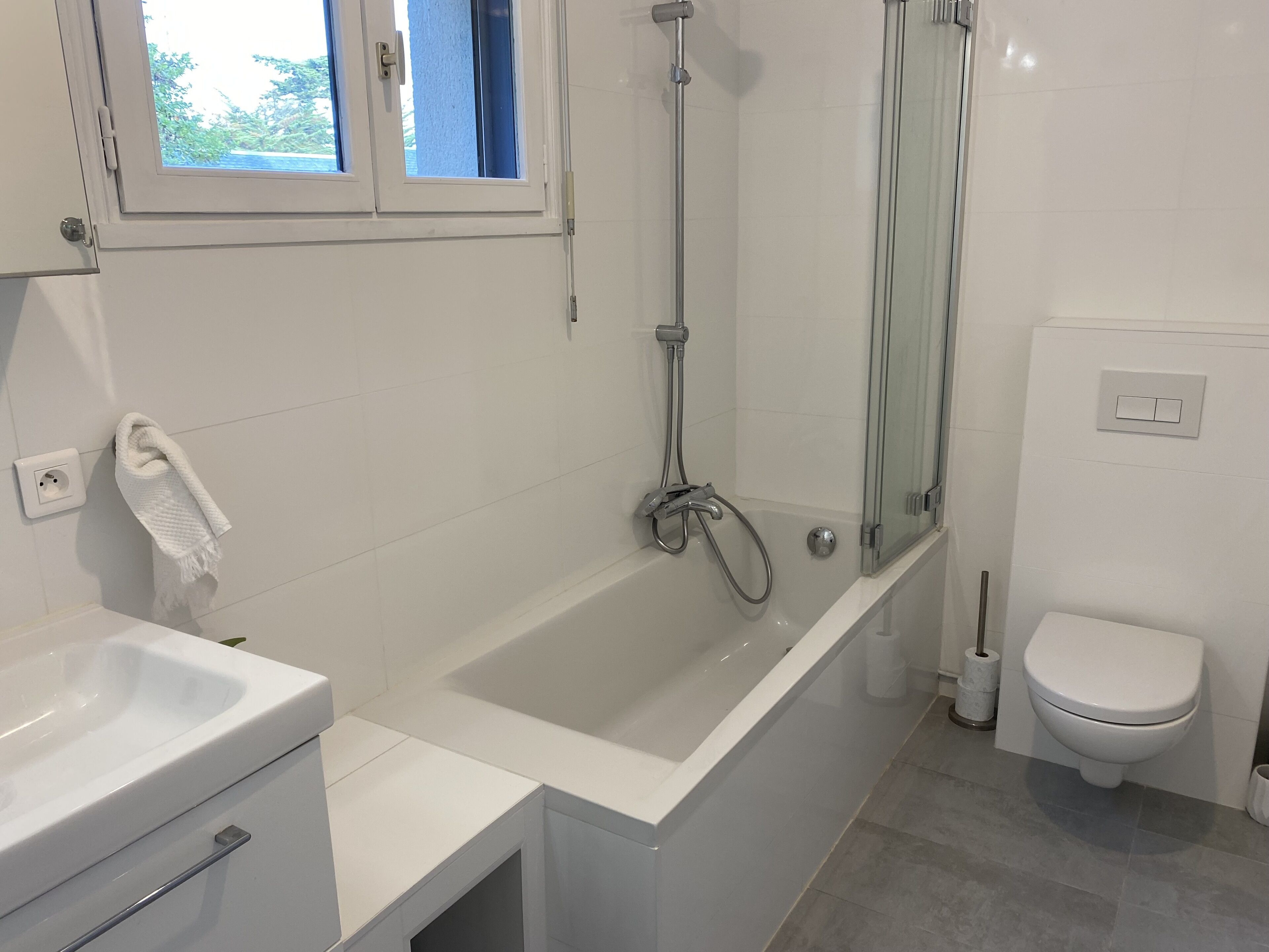 Une salle de bain moderne équipée d’une baignoire, d’une douche, de toilettes et d’une fenêtre donnant sur la verdure.