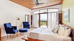 Ein geräumiges Schlafzimmer mit einem großen Bett, zwei blauen Sesseln, einer Holzbalkendecke und einem Balkon mit Blick.
