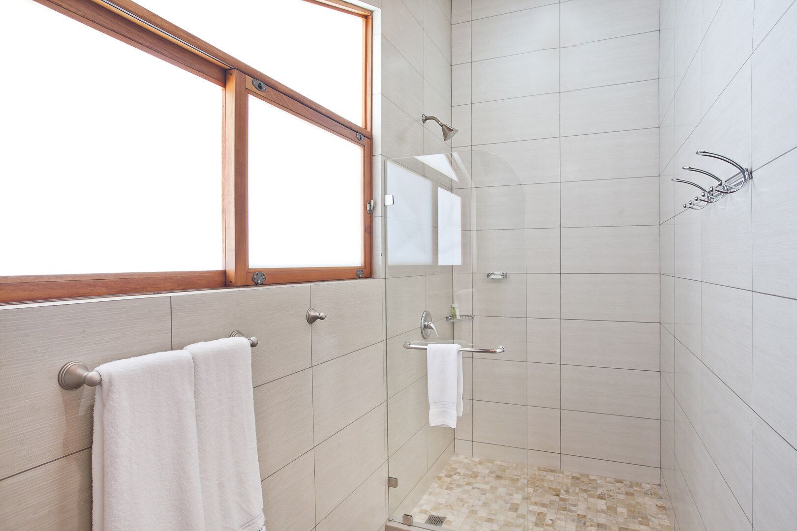 A modern bathroom with a glass shower enclosure, a wooden-framed window, and a tiled floor.