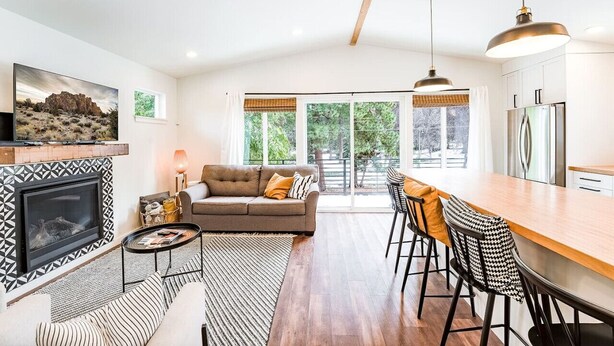 A modern kitchen with a large island, a dining area with bar stools, a fireplace, and a large window with a view of greenery. - Midtown gem I Park view I Full kitchen (Bend)