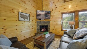 A cozy cabin interior with a fireplace, wooden walls, a mounted TV, and a comfortable seating area.