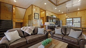 L’intérieur chaleureux d’un chalet, avec un revêtement mural en bois, un ensemble de canapés, un coin cuisine et une fenêtre équipée de stores.