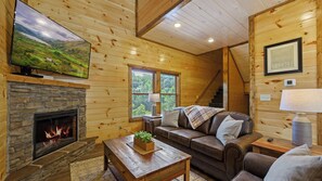 L’intérieur chaleureux d’un chalet avec une cheminée en pierre, un canapé en cuir, une table basse en bois et un téléviseur fixé au mur.