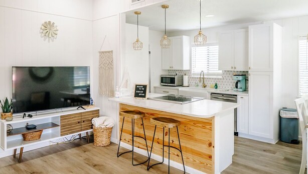 A modern kitchen with white cabinetry, a large island with stools, a flat-screen TV, and pendant lighting. - Riverfront Cottage Retreat with 2 Docks + Scenic Views on the St. John’s River! (Satsuma)
