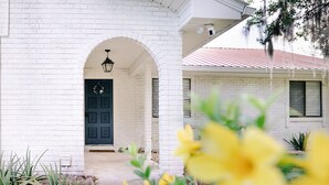 A white brick house with a dark blue door, a covered porch, and a small garden with yellow flowers. - Riverfront Cottage Retreat with 2 Docks + Scenic Views on the St. John’s River! (Satsuma)