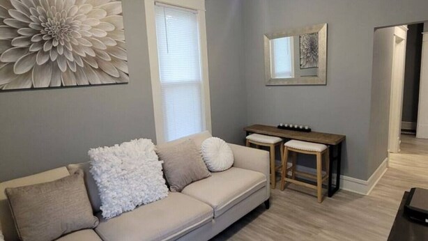 Ett vardagsrum med en beige soffa, två vita kuddar, ett träbord och ett stort blommönstrat konstverk på väggen.