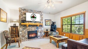 A cozy living room with a stone fireplace, a bear statue, and a large window with a view of trees.
