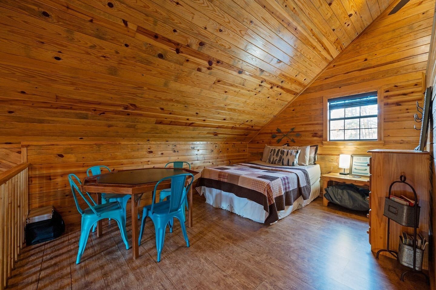 Een knus houten kamer met een houten plafond, een bed met quilt, een tafel met stoelen en een raam.