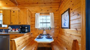 Ein Holzblockhaus-Interieur mit Küchenbereich, einem Esstisch für vier Personen und einem Fenster mit Vorhängen.