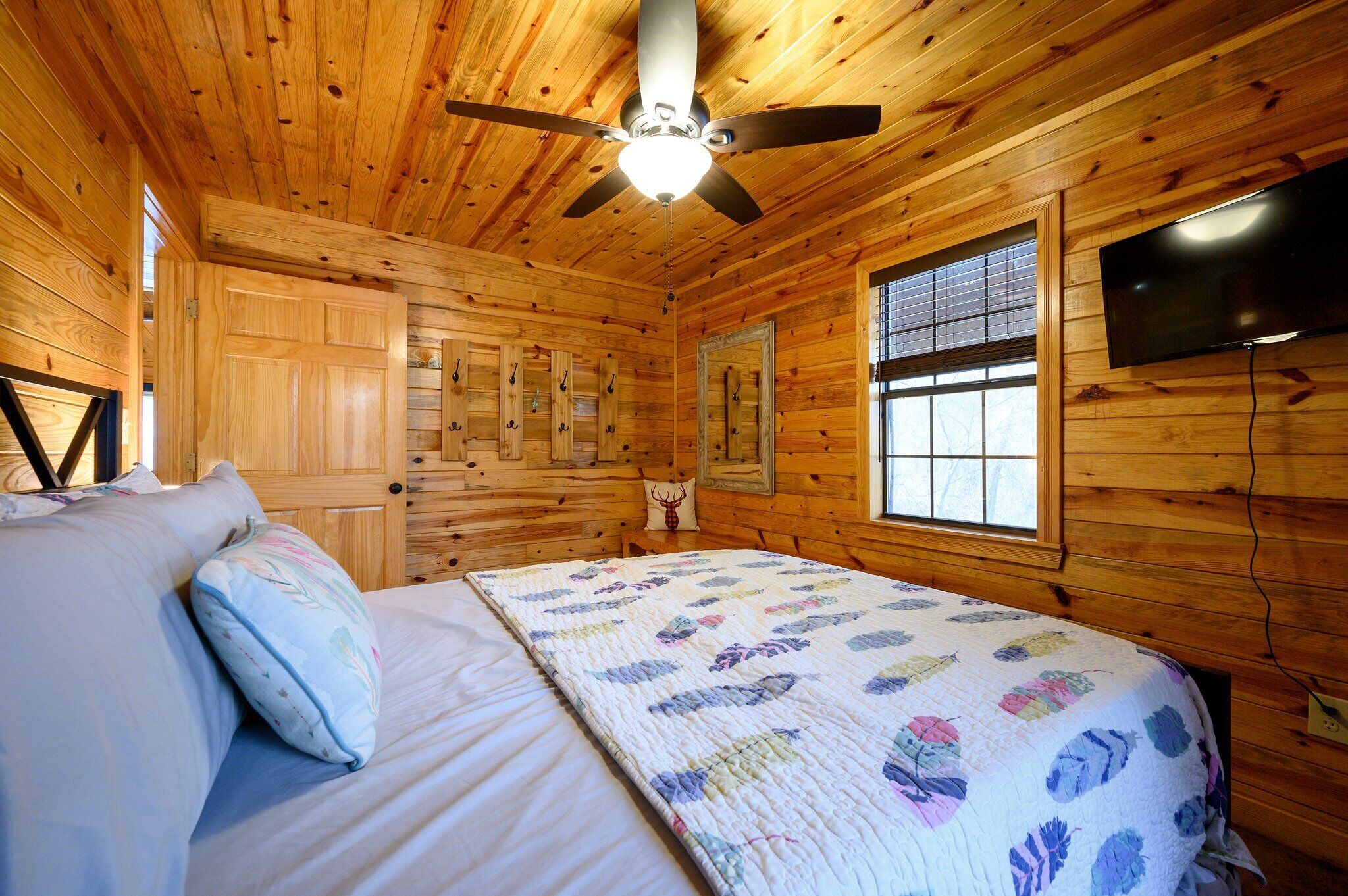A wooden cabin bedroom with a bed, pillows, a ceiling fan, a television, and a window.