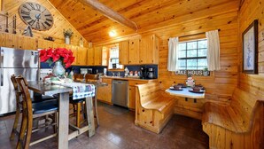 Ein rustikales Blockhaus-Interieur mit Holzwänden, einer Küchenzeile, einem Esstisch mit Stühlen, einer Bank und einer Wanduhr.