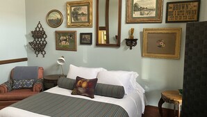 A neatly arranged bedroom with a large bed, a chair, a small table, and various framed pictures on the wall.