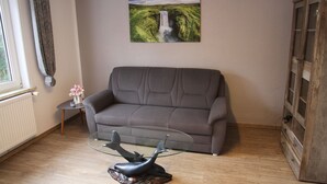 A living room with a grey sofa, a glass coffee table, a decorative fish sculpture, a painting of a waterfall, a wooden cabinet, and a radiator. - Apartment Julius 3 - nice, quiet and right in the middle - (Sankt Andreasberg)